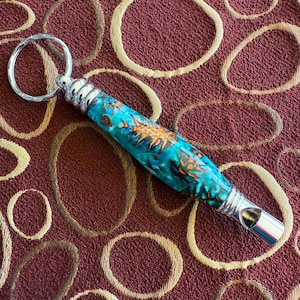 May include: A handcrafted whistle with a vibrant turquoise and orange resin body, accented with silver-toned metal components. The whistle includes a key ring attachment. The background is a textured maroon fabric with a circular pattern.
