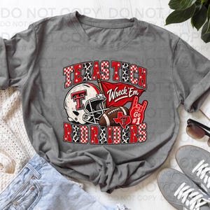 Op de afbeelding: Een grijs t-shirt met een afbeelding van een witte footballhelm, een bruine football, een rode vlag met de tekst Wreck'Em en een rood handgebaar met de tekst Go #1. De tekst Texas Tech Red Raiders is weergegeven in rode en witte patronen.