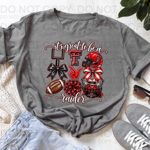 Könnte beinhalten: Ein anthrazitfarbenes Kurzarm-T-Shirt mit einem Grafikdruck und dem Text "it's great to be a raider". Das Design zeigt ein Football-Tor, ein rotes "TT"-Logo, einen Football-Helm, eine schwarze Schleife, einen roten Schaumstofffinger und ein Maskottchen.