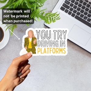 May include: A white sticker with the text "YOU TRY DRIVING IN PLATFORMS" in black and yellow. The sticker features a cartoon illustration of a woman in a yellow plaid suit. The sticker is held by a hand.