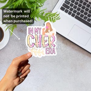 May include: A sticker with the text "IN MY CHER ERA" in pink and yellow gradient letters, featuring a cartoon illustration of a woman holding a makeup applicator. The sticker has a white border and a floral design at the bottom.