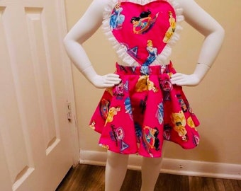 Puede incluir: Un conjunto de dos piezas de color rosa brillante que presenta un corpiño en forma de corazón con ribete de volantes blancos y una falda acampanada a juego. La tela está estampada con varios personajes de princesas y formas geométricas.