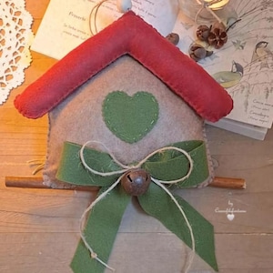 Può includere: Un ornamento per la casa in feltro con un tetto rosso, un cuore verde e un fiocco verde con un campanello. L'ornamento è appeso a un bastoncino di legno.