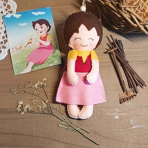 Può includere: Una bambola di feltro fatta a mano con capelli castani, un vestito rosa e una camicia gialla. La bambola sorride e ha le mani giunte davanti a sé. La bambola è seduta su una superficie di legno.