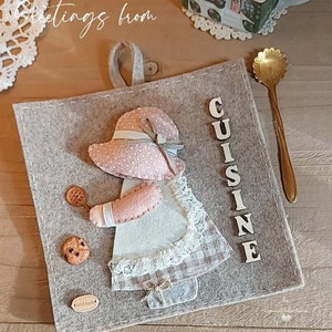 Può includere: Un appendiabiti da cucina in feltro grigio con una bambola di feltro che tiene un biscotto e una spatola. La bambola indossa un vestito rosa e bianco con un bordo di pizzo bianco. La parola "CUISINE" è scritta in lettere bianche sull'appendiabiti. L'appendiabiti ha anche un piccolo cartellino marrone che dice "fatto a mano".
