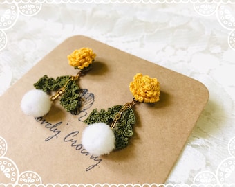 May include: A pair of crocheted earrings with yellow flower accents and white pom-poms. The earrings are hanging from gold chains and are displayed on a brown card with the text "Lovely Crown".
