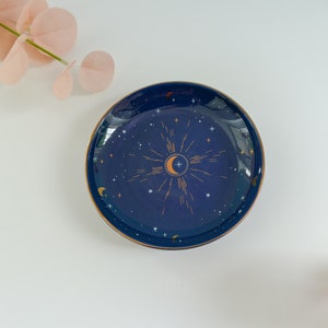 May include: A blue ceramic trinket dish with a gold rim and a crescent moon design with stars and a sunburst pattern.