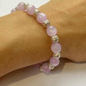 May include: A light purple beaded bracelet with silver filigree spacers. The bracelet, worn on a wrist, features round, polished beads in a soft pastel hue. The beads are separated by silver-toned metal.