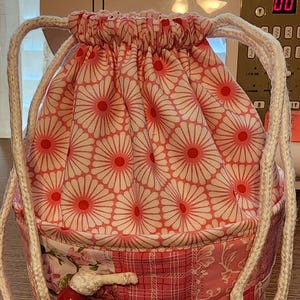 May include: A handmade drawstring bag with a floral pattern in shades of pink and white. The bag features a patchwork design on the lower half and is accented with white rope handles and red wooden beads.