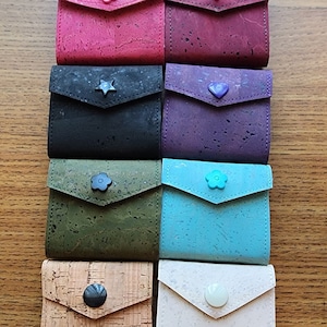 May include: Eight small cork wallets in different colors with various button designs. The wallets are arranged in a row on a wooden surface.