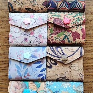 May include: Nine cork wallets with different patterns and colors. Each wallet has a button closure with a different design. The wallets are arranged in three rows of three.