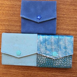 May include: Three small, square wallets in shades of blue and teal are displayed on a wooden surface. The top wallet is a solid blue with a star-shaped button. The other two have floral and textured details.