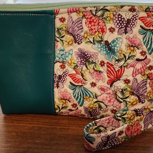 May include: A teal zippered pouch with a white fabric flap featuring a colorful butterfly print. The pouch has a detachable strap with a silver clasp.