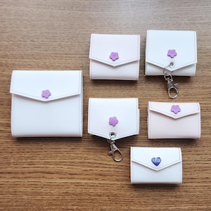 May include: A collection of small, white and pink leather wallets with a flower or heart-shaped button closure. Some wallets have a silver-colored clasp. The wallets are arranged on a wooden surface.