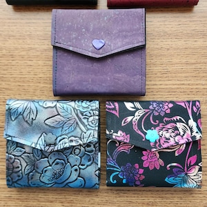 May include: Five square wallets with flap closures and snap buttons. The top row features solid black and dark red cork textures. The middle row is a solid purple cork wallet. The bottom row includes a blue floral pattern and a black floral print.