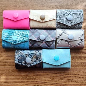 May include: Eight small leather wallets with different patterns and colors. The wallets have a single button closure. The wallets are arranged in two rows of four.