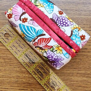 May include: A rectangular fabric case with a butterfly pattern in pink, blue, and purple, accented with red trim. A clear ruler with yellow markings is next to the case. The ruler is marked in inches.