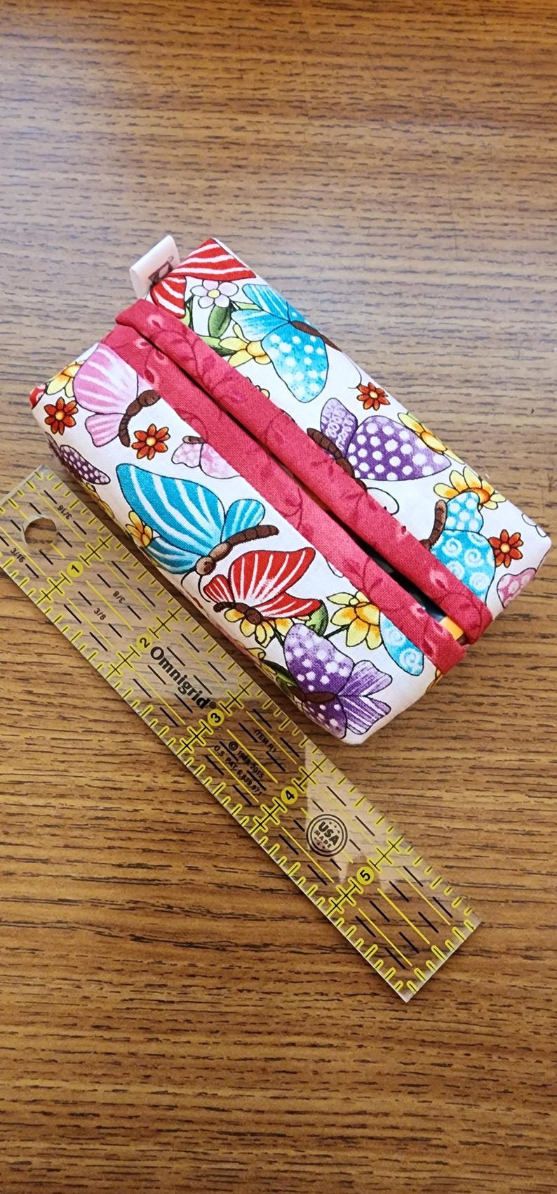 Op de afbeelding: Een rechthoekige stoffen etui met een vlinderpatroon in roze, blauw en paars, geaccentueerd met rode bies. Een doorzichtige liniaal met gele markeringen ligt naast de etui. De liniaal is gemarkeerd in centimeters.