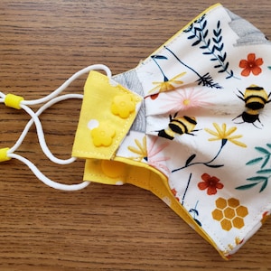 May include: A white face mask with a floral and bee print. The mask has yellow adjustable straps with yellow buttons. The mask is made of fabric and has a pleated design.