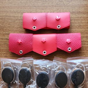 May include: Five red leather keychains with different colored heart, star, and flower designs. Each keychain is attached to a black retractable badge holder.