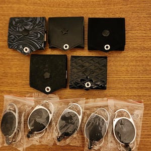 May include: Six black leather card holders with different designs and six silver retractable badge holders in clear plastic packaging.