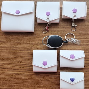 May include: Six white zippered pouches with purple flower and heart embellishments. The pouches are different sizes and some have keychains attached.