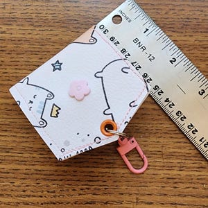 May include: A small, light pink cardholder with a cartoon design, a pink flower embellishment, and a pink clasp. A ruler is placed diagonally across the cardholder, measuring in inches.