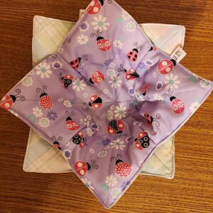May include: Two handmade bowl cozies. The top cozy is lavender with a ladybug and floral pattern. The bottom cozy is light green with a plaid pattern. Both are square and sit on a wooden surface.