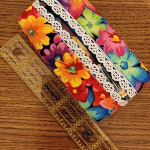 May include: A floral fabric item with a white lace trim, possibly a pencil case or glasses case, is next to a clear ruler. The fabric features colorful flowers in shades of orange, yellow, pink, and blue.