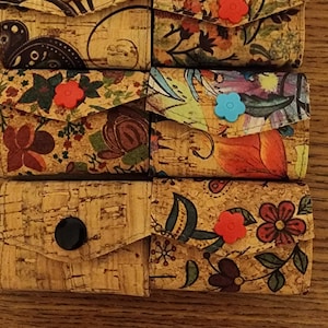 May include: Six small cork wallets with different floral patterns. Each wallet has a button closure and a flap. The wallets are arranged in a stack.