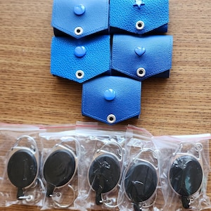 May include: Six blue leather keychains with different colored buttons and a star, heart, and circle design. Five black retractable keychains in individual plastic bags.