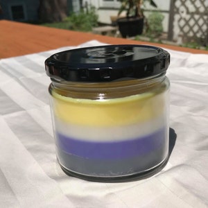 May include: A layered candle in a glass jar with a black lid. The candle is yellow, white, purple, and gray.