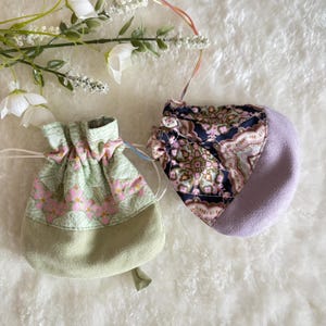 May include: Two small drawstring pouches. One is light green with pink floral patterns, the other is a mix of patterned fabric and lavender. Both have drawstrings for closure. The pouches are on a white, textured surface.