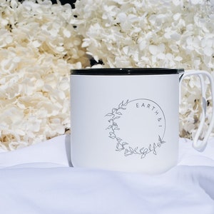 May include: White enamel camping mug with a black rim and handle. The mug has a black and white logo that says "Earth & I" with a circle of leaves around it.