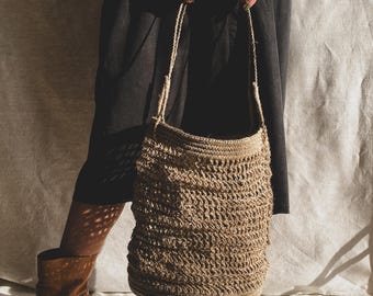 May include: A large, brown, crocheted tote bag with a thick rope strap. The bag is being held by a person wearing a black dress and brown boots.