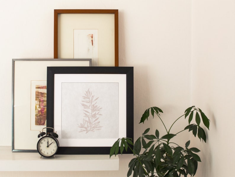 May include: Three framed pictures on a white shelf with a black alarm clock in front of them. The picture on the left has a colorful abstract design. The middle picture has a black frame and a white background with a delicate, light pink leaf design. The picture on the right has a brown frame and a white background with a faint, light pink design.