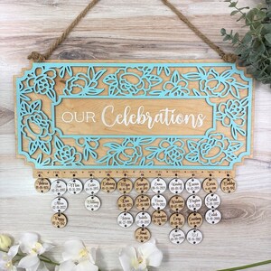 May include: A wooden sign with a light blue floral design and the text "OUR Celebrations". The sign has a rope hanger and is decorated with small wooden circles with names and dates.