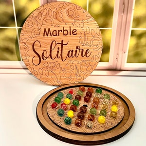 May include: A wooden marble solitaire game with a circular board and a separate lid. The lid has a floral design and the words "Marble Solitaire" engraved on it. The board has a similar floral design and is filled with colorful marbles.