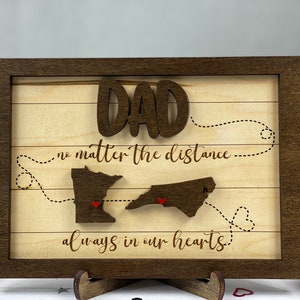 Puede incluir: Cartel enmarcado de madera con la palabra "DAD" en letras en relieve. El cartel presenta dos contornos de estados con corazones, conectados por líneas de puntos, y las frases "no matter the distance" y "always in our hearts."