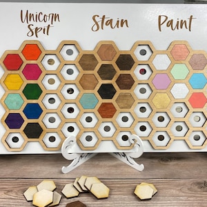 May include: A wooden board with a honeycomb pattern, displaying various colors of paint and stain samples. The board is labeled "Unicorn Spit", "Stain", and "Paint".