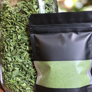 May include: A clear glass jar filled with dried green leaves, next to a black pouch containing a fine, green powder. The leaves appear to be a dried herb, and the powder may be a spice or supplement.