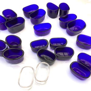 May include: A collection of oval-shaped glass dishes in a vibrant cobalt blue. The dishes have a dark rim and are arranged on a white surface. Two clear glass dishes are also present. These could be used for serving dips or small portions.