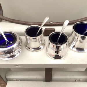 May include: A set of four silver salt cellars, each with a cobalt blue glass liner and a small silver spoon. The cellars vary in shape, including a decorative round design and cylindrical forms. They are displayed on a white surface.