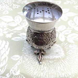 Könnte beinhalten: Ein silberfarbener, verzierter Tee-Infuser mit perforiertem Deckel und einem detaillierten, distelartigen Sockel. Der Infuser steht auf drei dekorativen Füßen. Der Hintergrund ist eine hellgrüne Tischdecke mit floralem Muster. Ca. 10 cm hoch.