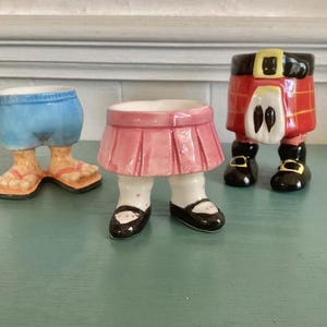 May include: Three ceramic egg cups shaped like lower bodies. One wears blue shorts with flip-flops, one wears a pink pleated skirt with black shoes, and one wears a red tartan kilt with a sporran and black buckled shoes. Each cup has a hollow top.