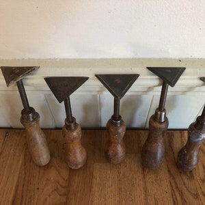 Könnte beinhalten: Sechs antike Spachtel aus Metall mit Holzgriffen. Die Spachtel haben dreieckige Klingen und sind in einer Reihe auf einer Holzoberfläche angeordnet.