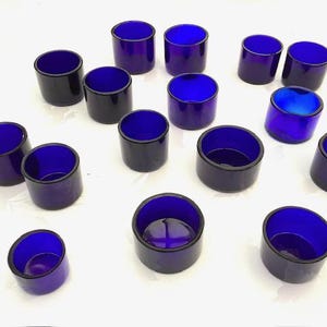 Peut inclure: Une collection de bougeoirs votifs en verre bleu cobalt. Les supports cylindriques ont un extérieur sombre et un intérieur bleu vif. Les dessus ouverts et les bases arrondies sont visibles. Les bougeoirs sont disposés sur une surface blanche.