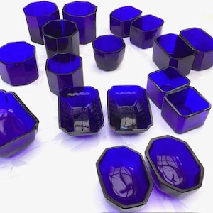 May include: An assortment of cobalt blue glass containers in various geometric shapes. The collection includes octagonal tumblers, rectangular dishes, and oval bowls, all with a glossy finish. The containers are arranged on a white surface.