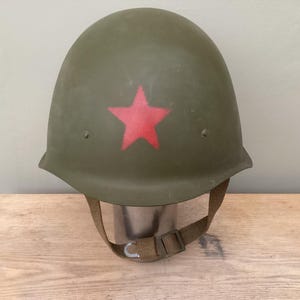 Puede incluir: Casco militar verde oliva con una estrella roja en la parte delantera. El casco tiene una correa para la barbilla y está sobre un soporte metálico. El casco es de metal.