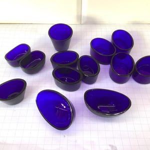 Könnte beinhalten: Eine Auswahl an kobaltblauen Glasschalen und -bechern. Die Schalen sind oval mit einem dunklen Rand, während die Becher zylindrisch sind. Die Objekte sind auf einer weißen Oberfläche mit einem Rastermuster angeordnet. Das Glas hat eine glänzende Oberfläche.